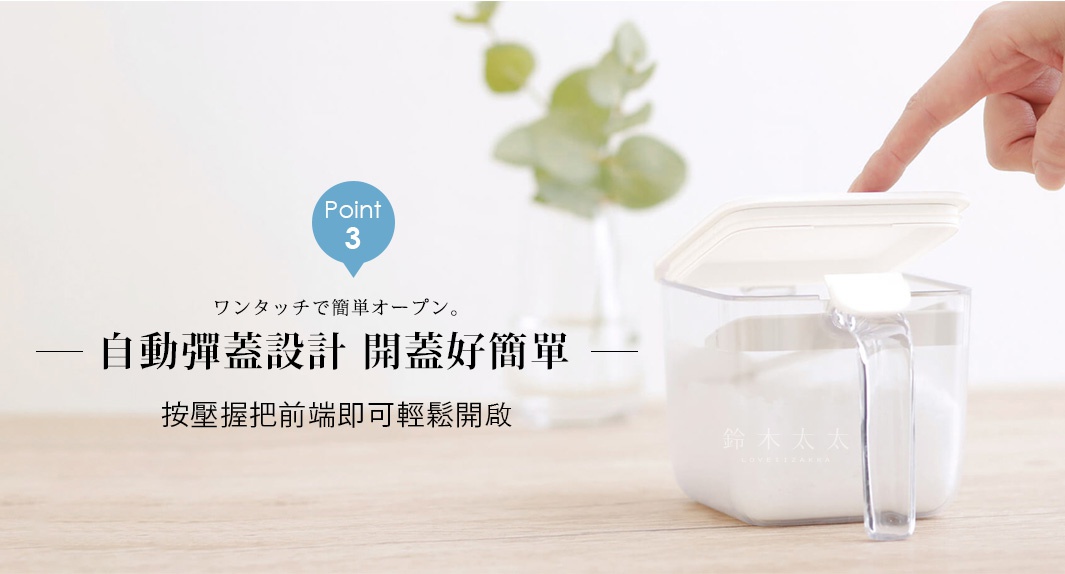 透明材質的密封調味料盒放在木質桌面上，右側有人手輕壓把手前端的開關，上蓋呈現自動彈開狀態，盒身內裝有白色調味料，背景為模糊的綠色植栽，強調單手即可輕鬆開啟的自動彈蓋設計。