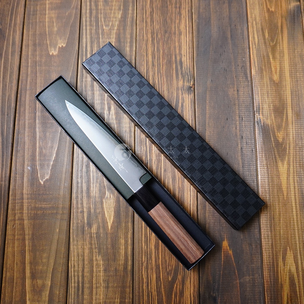 [景清] Chromax鋼水果刀，刀身呈銀白色，握柄為胡桃木材質，刀刃長度約15公分，置於黑色禮盒內，禮盒表面有格子紋路，整體擺放在木紋桌面上。