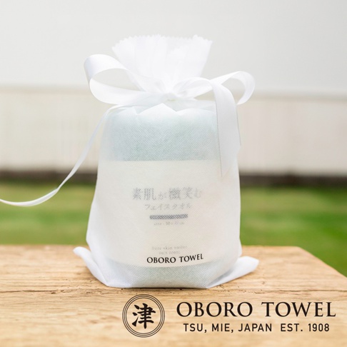 白色束口袋包裝著綠色與白色相間的毛巾，束口袋頂部繫著白色緞帶，袋身印有日文「素肌が微笑む フェイスタオル」及英文「OBORO TOWEL」字樣。