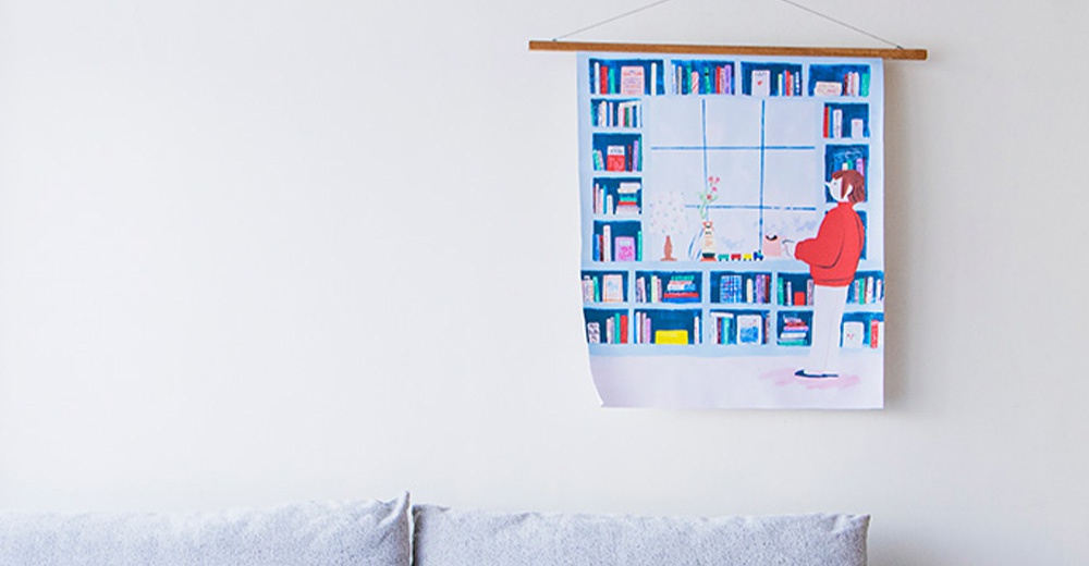 無框帆布藝術海報，描繪一幅柔和色調的室內場景，左側為白色牆面，右側懸掛著藍色調的帆布藝術畫。畫中以簡約線條和色彩，呈現滿滿書架的背景，書架前有一位身穿紅色毛衣、手持杯子的年輕人，畫面風格溫馨。