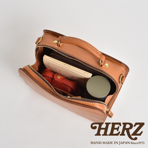 職人手作皮革兩用方形單肩包，淺棕色，拉鍊開啟，內有書本、紅色皮夾和圓形物體，搭配品牌標誌。