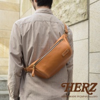 職人手作淺棕色皮革輕便斜背包，側面為圓潤弧形，頂部配有黃銅色拉鍊，右側有品牌皮標，側邊附有可調整的皮革揹帶。