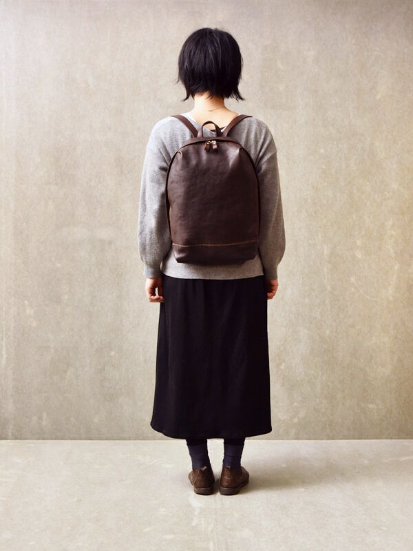 一位女士身後背著一個深棕色皮革後背包，背包呈簡約褶皺風，配有雙向拉鍊和一個較小的底部隔層。