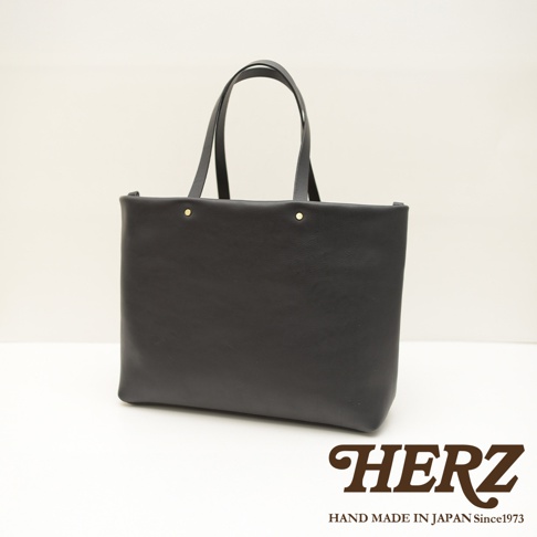 黑色皮革極簡托特包，配有兩條皮革提把，包身正面有兩個金色鉚釘裝飾，展現皮革的自然紋理。