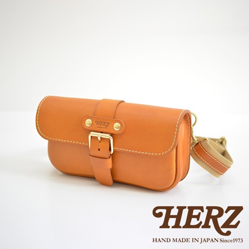 一個經典斜背包，採用 tan 色皮革製成，帶有單扣閉合設計，金色金屬配件，以及米色織帶。