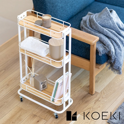 白色三層置物推車，附有木紋層板，架體為金屬材質，配有四個輪子。推車上擺放有毛巾、蠟燭、收納盒、擴香瓶及書籍。