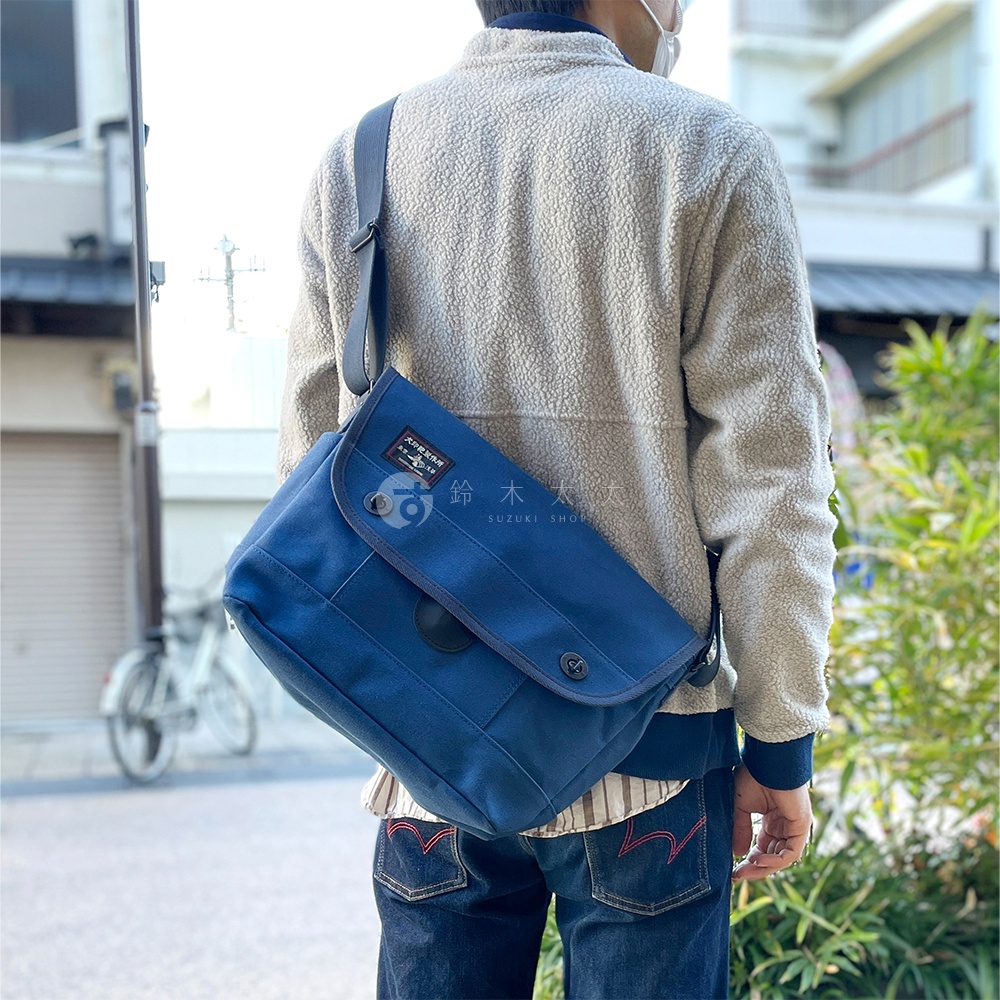 藍色純棉帆布雙扣郵差包，斜挎於身，包身設計簡潔，有明顯的縫線和金屬雙扣。