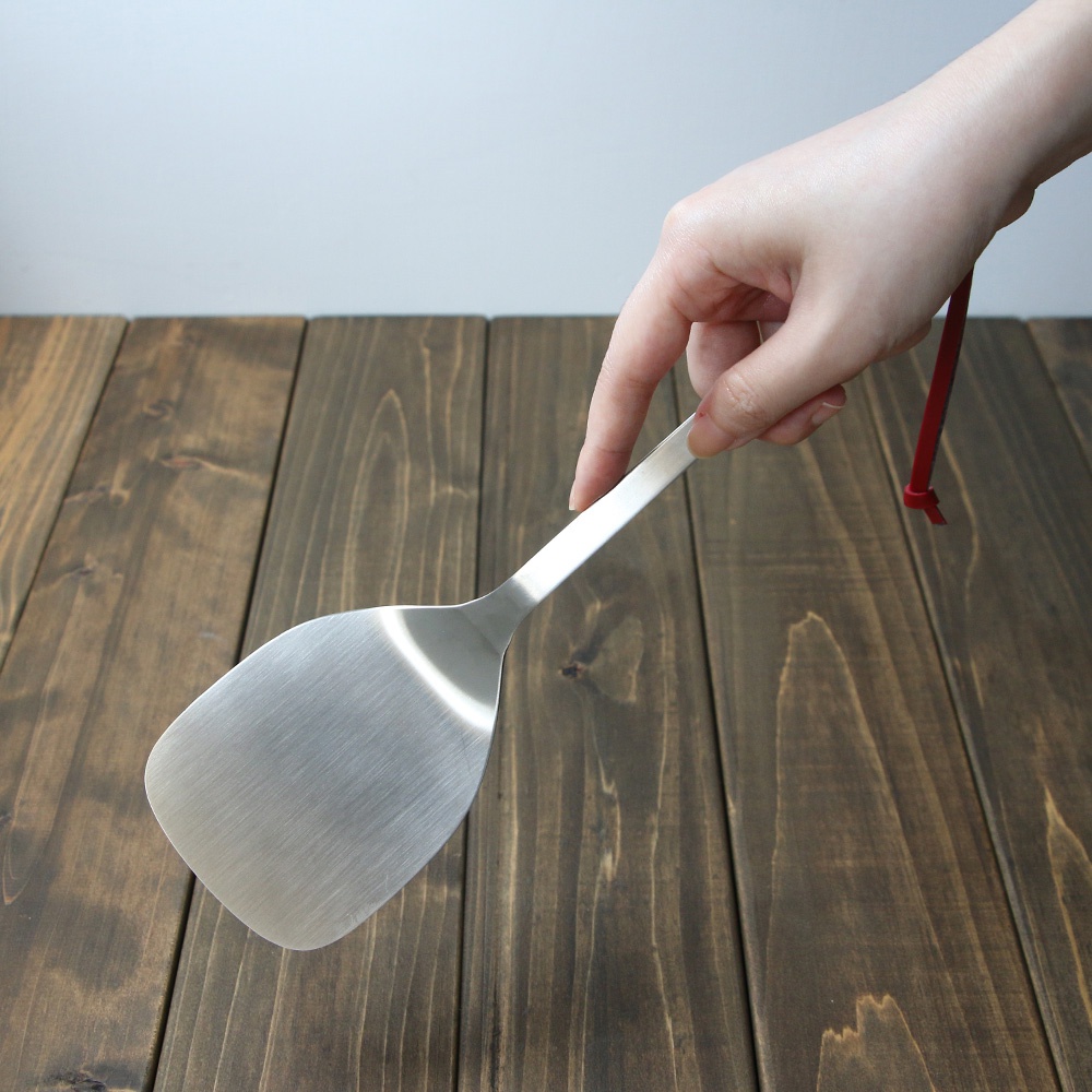 手持不鏽鋼萬用鍋鏟，鍋鏟呈銀白色，光滑表面，鍋鏟頭為寬扁扇形，手柄細長，末端有紅色掛繩。
