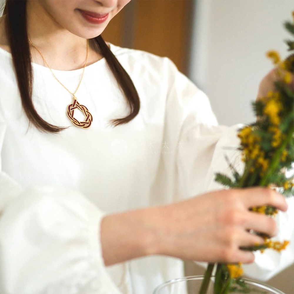 女性佩戴一條金色細鍊，鍊墜為紅色與金色的水引花結造型，簡約別緻。