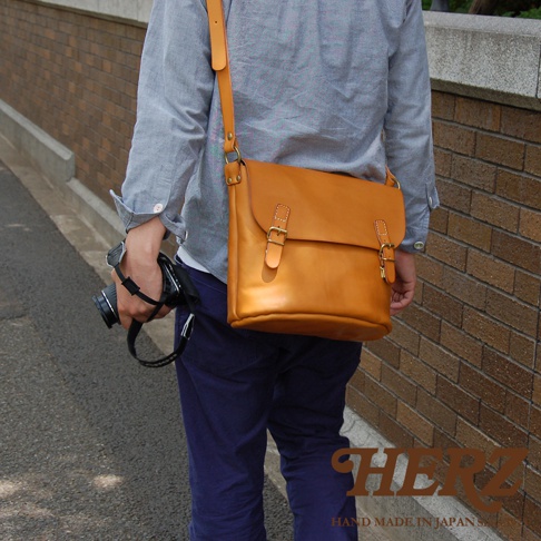 HERZ 職人手作皮革雙扣旅行郵差包，以焦糖色皮革製成，搭配斜揹帶與雙插釦設計，並附有相機。