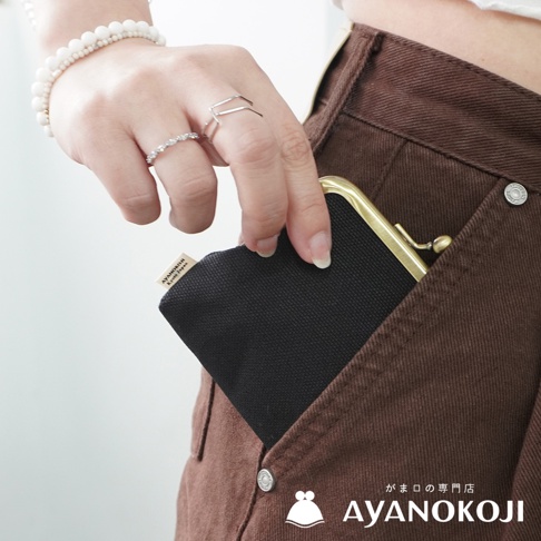 一位女子將一個黑色綾織布製的口金零錢包，從深棕色褲子的口袋中取出，零錢包的金屬夾閃閃發亮，背景可見女子手腕上的白色串珠手鍊和戒指，圖片右下角顯示AYANOKOJI品牌標誌。