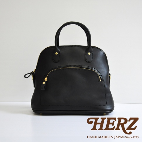 黑色皮革手提包，圓潤貝殼造型，頂部提把，正面設有拉鍊口袋，金色拉鍊與釦飾，手工製作。