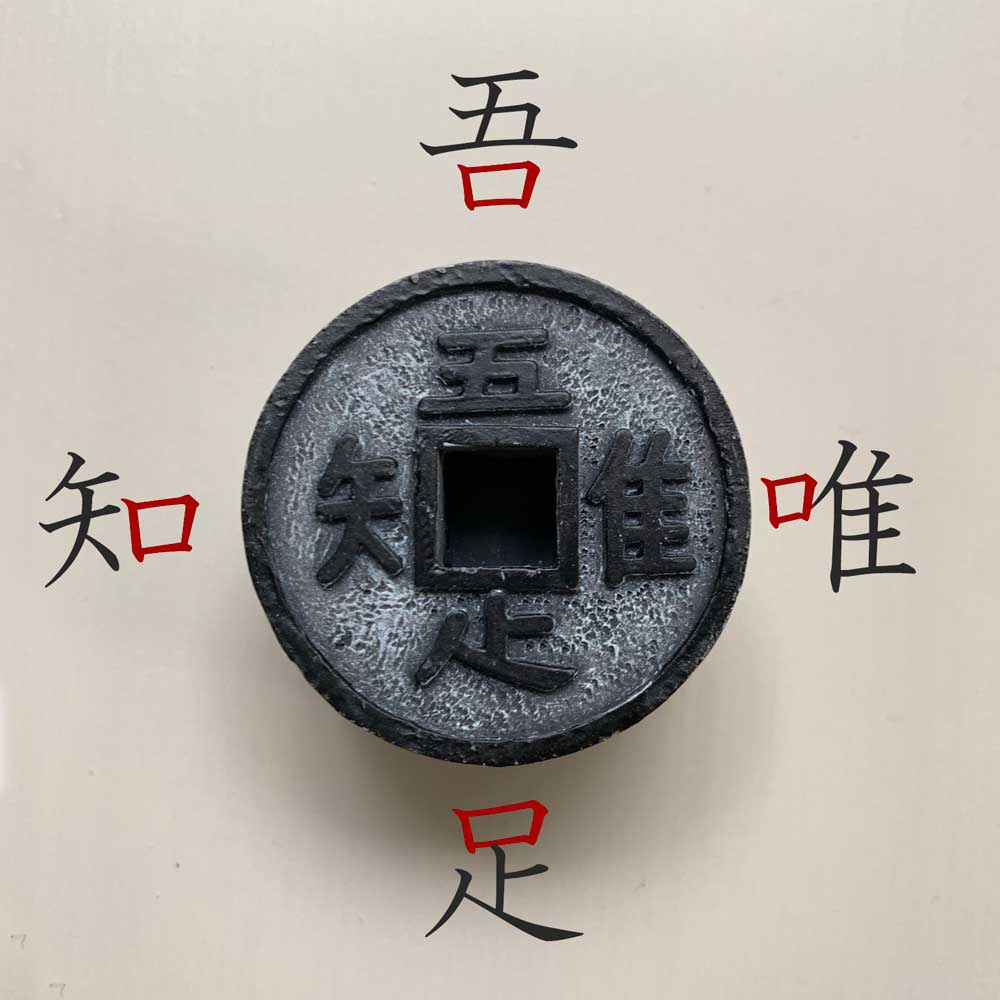 一個帶有方形開孔的圓形黑色金屬模型，表面有粗糙紋理，周圍環繞著黑色的中文「吾」、「知」、「足」、「唯」字樣，其中「吾」和「唯」的紅色部分點綴。