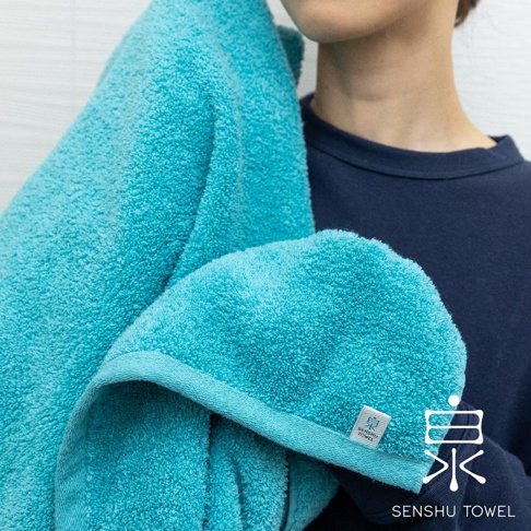 一件藍綠色長纖浴巾，材質柔軟蓬鬆，邊緣有細緻的縫線，浴巾上印有「SENSHU TOWEL」的白色小標籤。