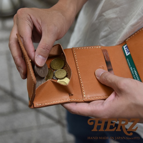 一個人手持日本HERZ手工製作的原色牛皮三折短夾，短夾展開露出設計寬敞的盒型零錢袋與放入其中的硬幣，右側皮夾夾層中插有一張三井住友VISA信用卡，整體呈現出職人皮革工藝的細膩質感。