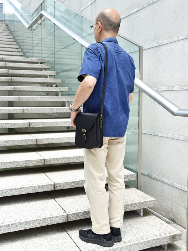 一名男性穿著藍色短袖襯衫和卡其色長褲，斜背著黑色皮革翻蓋迷你單肩包，包包上有金色扣環。