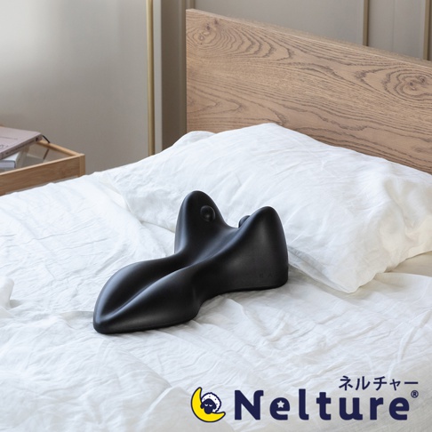 黑色肩頸背拉伸牽引枕，呈現人體工學的特殊造型，置於白色床單和枕頭上，背景為木質床頭板。