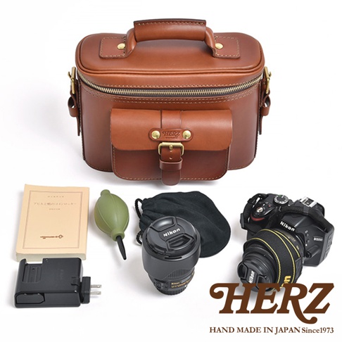 日本 HERZ 職人手作皮革厚實相機包 N-8，搭配 Nikon 相機、鏡頭、相機清潔組、充電器與日文書籍，呈現豐富的攝影配件組合，HERZ 品牌 LOGO 標示於包包正面。