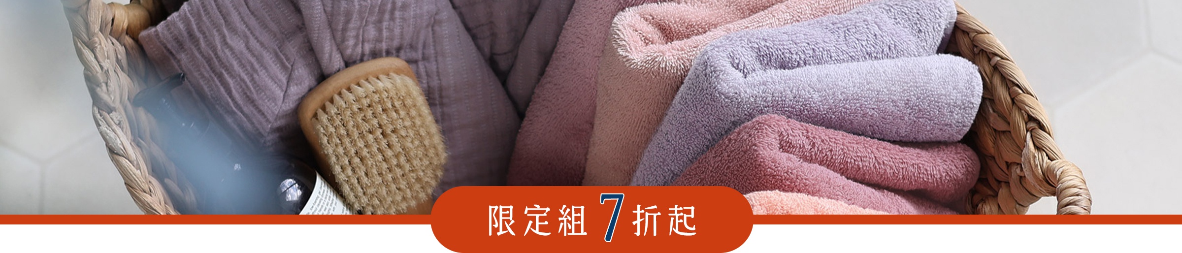 藤編籃內擺放著柔軟的粉色與紫色毛巾，旁邊放置一支木質沐浴刷與深色瓶裝用品，籃子下方標示限定組合七折起。