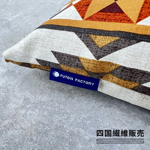 這是一款具有幾何圖形的民族風枕頭，枕套採用米白色基底搭配橙色、深褐色與芥末黃色的三角幾何花紋，枕頭邊緣縫有一個深藍色的方形標籤，上面印有白色 FUTON FACTORY 字樣，整體風格簡約且充滿異國情調，右下角標註有四國纖維販售字樣。