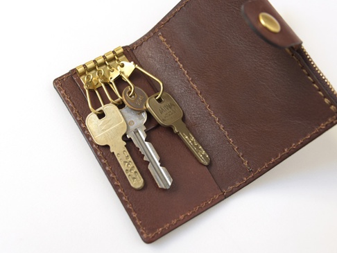 深棕色皮革製的六鉤鑰匙包，配有黃銅色拉鍊及按扣，內含數把鑰匙，展示了精緻的縫線工藝。