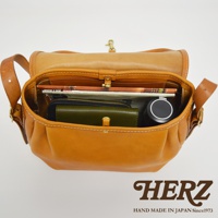 職人手作皮革復古翻蓋郵差包，可見內部裝有雜誌、綠色皮夾、黑色圓筒狀物品，包身為淺棕色皮革，縫線明顯。