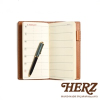 職人手作皮革記事本書套，淺棕色皮革外觀，內含週曆記事本，書頁上印有日期與時間格線，一支綠色與金色筆夾的鋼筆置於書本中央。