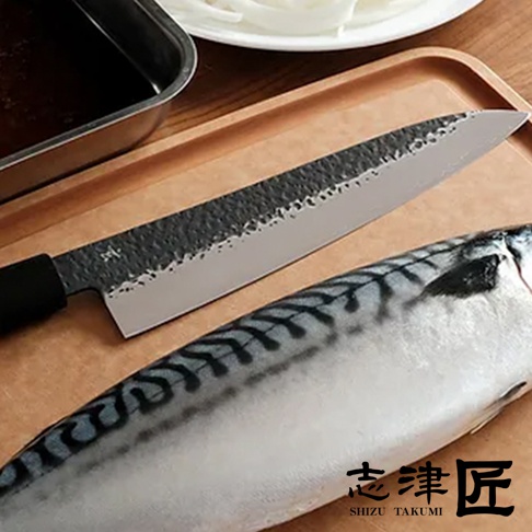 玄 / Chef's knife 主廚刀18cm，鋒利的銀色刀刃，刀身帶有獨特的黑色錘紋和細緻的紋理，搭配黑色刀柄，置於木質砧板上，旁邊有一條新鮮的鯖魚。