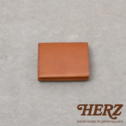 這是一款由日本HERZ品牌推出的手工皮革短夾，以義大利Minerva Liscio皮革製作，外觀呈現溫潤的棕色質感，採用極簡的長幅單片皮革包覆設計，側面可見細緻的職人手縫線跡，整體造型小巧俐落。