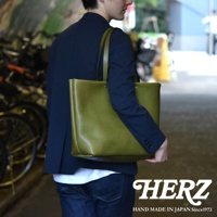 橄欖綠皮革極簡托特包，配有兩條肩帶，包包正面有兩顆鉚釘裝飾，由一位穿著海軍藍西裝外套和牛仔褲的男士攜帶。