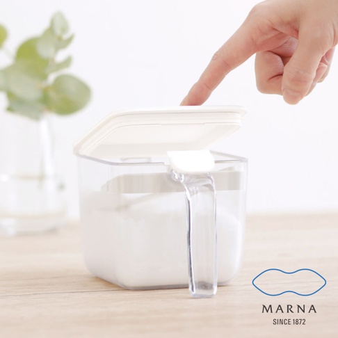 一隻手正輕觸一個透明的方形調味料盒，盒內裝有白色粉末，蓋子呈現半開狀態，旁邊擺放著一株綠色植物，右下角印有 MARNA SINCE 1872 的品牌標誌。