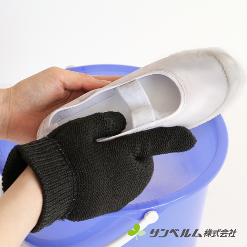 一隻黑色編織手套戴在手上，正在清洗一隻白色織布材質的運動鞋，運動鞋上有白色魔鬼氈黏帶，置於藍色水桶中。