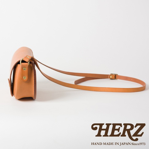 職人手作皮革迷你馬鞍包，展示的是一個卡其色皮革迷你馬鞍包，側面視角，附有細長皮革肩帶，肩帶上有金屬調節扣。