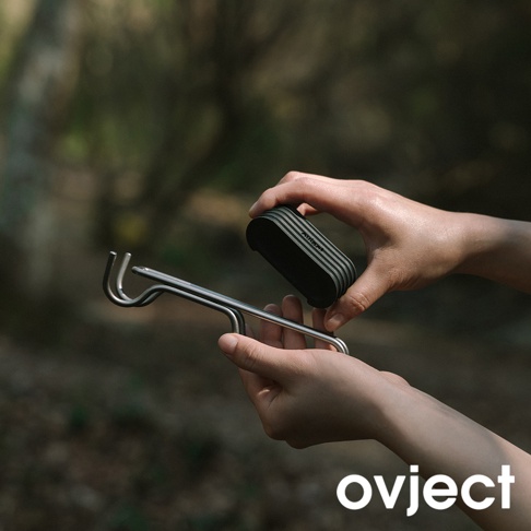 雙手持著銀色金屬製成的鍋夾，夾頭呈彎鉤狀，另一手持著黑色帶有條紋的隔熱握把。