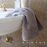 一條灰色極柔蓬鬆的今治毛巾，掛在白色洗手台上，背景是淺棕色紋理牆面和金色水龍頭。