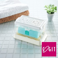 白色塑膠製的多功能免沾手泡沫洗衣皂盒，盒蓋上有通風孔。盒內有一個淺藍色的洗衣皂，置於黃色的刷毛底座上。背景是堆疊的毛巾和綠色植物。