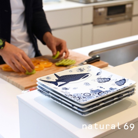 一疊白色方形餐盤，盤面繪有藍色海洋生物圖案，包括鯨魚、海龜和水母。背景為廚房場景，有人正在切水果。