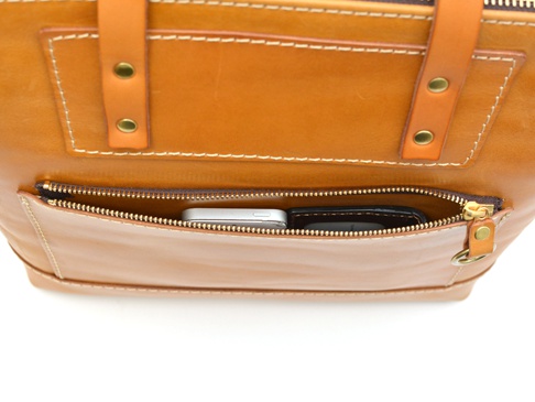 職人手作皮革威靈頓直式拉鍊托特包，採用光滑的棕褐色皮革製成，設有一個橫跨包身前方的拉鍊口袋，口袋內可見一部銀色手機和一個黑色錢包。包包有清晰的縫線細節和金屬釘。