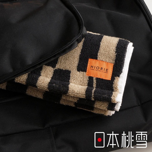 一條帶有黑色和米色格紋的毛巾，標有「HIORIE TOWEL FACTORY IN JAPAN」橙色標籤，放置於一個打開的黑色包內，右下角有「日本桃雪」的字樣。