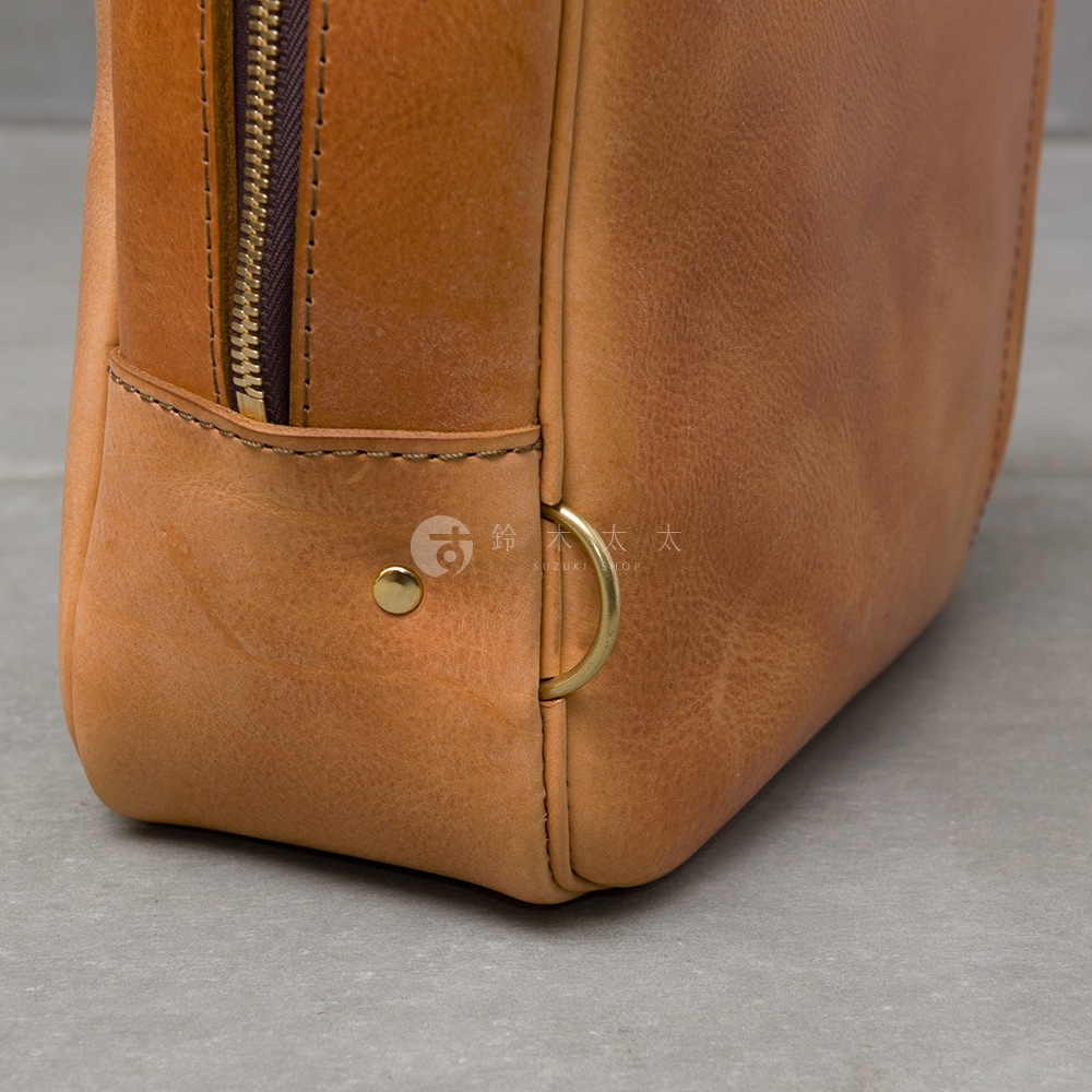 職人手作皮革Organ商務後背包的細節，包含金屬拉鍊、銅質铆釘和圓形金屬環，整體呈現溫暖的棕褐色調。