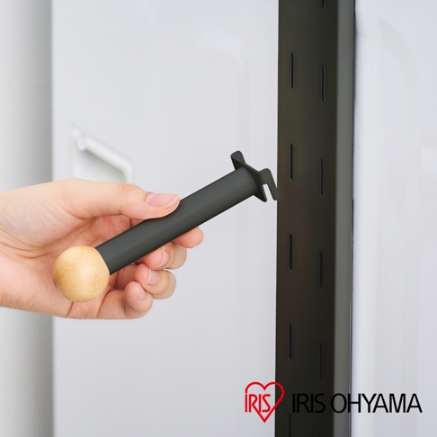 手持黑色圓柱形磁吸式洗衣機收納掛架，一端為木質圓球，另一端有黑色塑膠掛鉤，正要插入黑色金屬掛架的孔洞中。