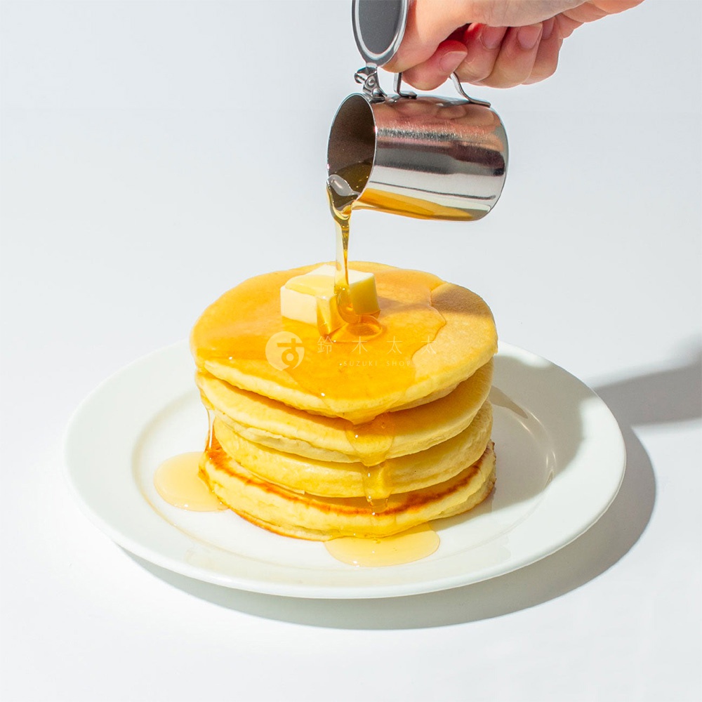 不鏽鋼附蓋蜂蜜盅，正在為堆疊的鬆餅淋上金黃色蜂蜜，鬆餅上放有一塊奶油。