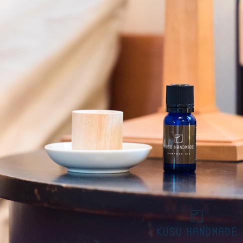 淺色木質擴香木筒置於白色小碟上，旁邊擺放著一瓶藍色小玻璃瓶裝的精油，瓶身貼有「KUSU HANDMADE CAMPHOR OIL」標籤。