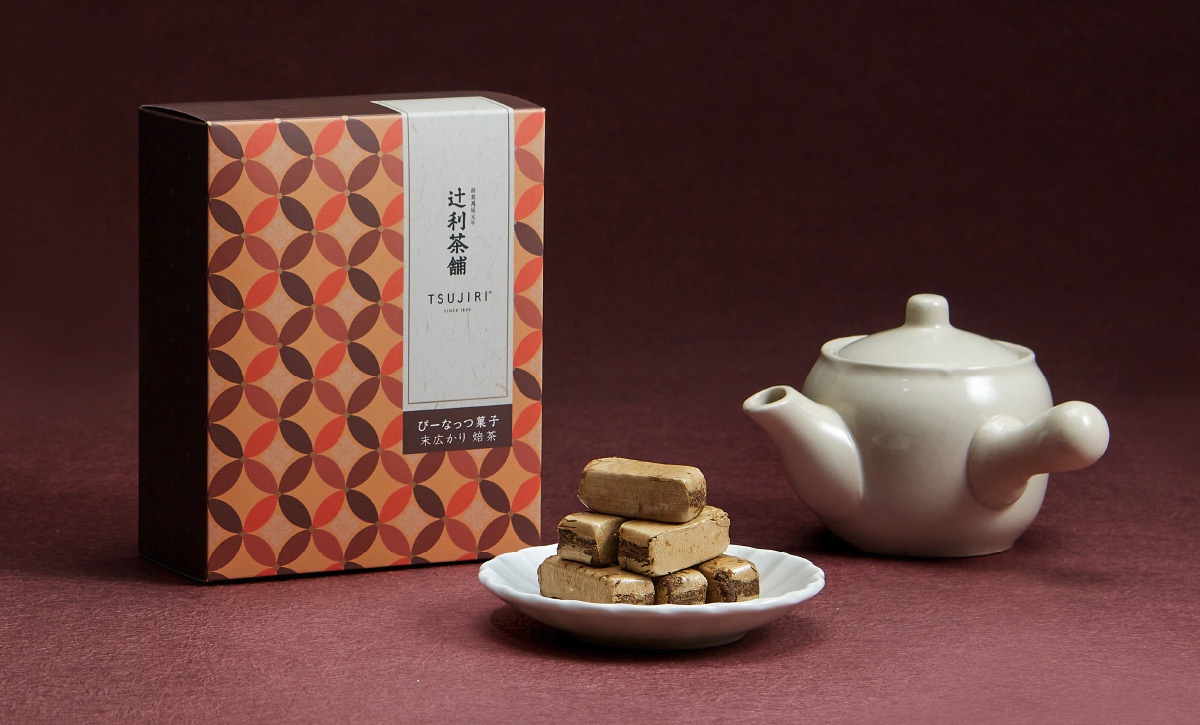辻利茶舖的伴手禮包裝盒，旁放置白色小茶壺與盛裝數塊長條狀烤茶酥的白色花邊盤，整體呈現溫暖的棕色系色調，包裝盒上有菱形圖案及日文品名與品牌標誌。