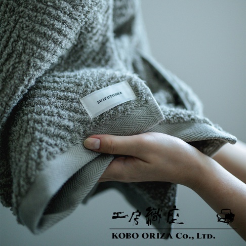 一條淺灰色人字紋有機棉毛巾，標籤印有「SUiFUTOSHA」，材質柔軟，觸感細緻。