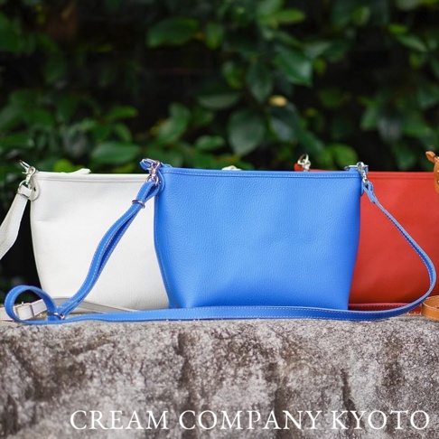 三款皮革隨身側背包，分別為白色、藍色和紅色，皆配有銀色金屬扣環和可調式皮革背帶，擺放在石塊上，背景為綠色植物。