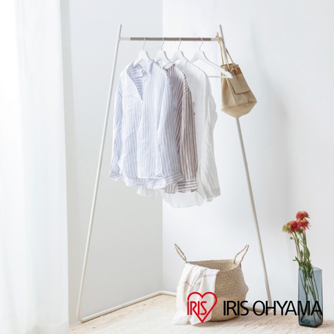 角落造型吊掛衣桿，白色金屬支架，懸掛多件條紋及素色襯衫，右側吊掛一個米色帆布袋，下方放置編織籃及鮮花。