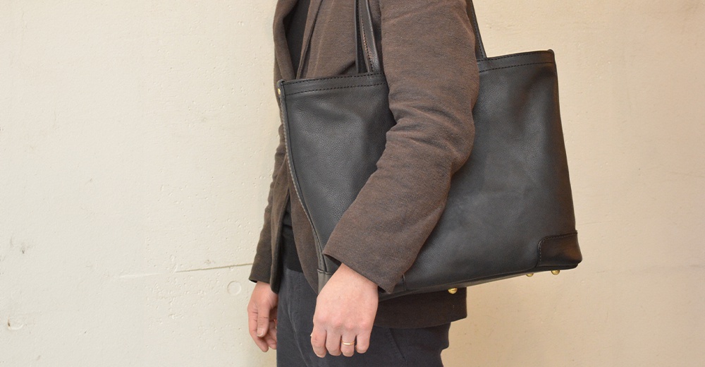 一個黑色皮革托特包，由一人肩背，包的側面可見縫線細節。