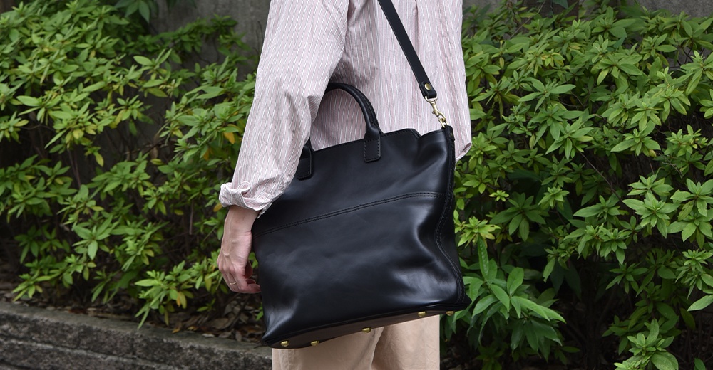 黑色皮革大容量托特包，配有提把和可拆卸肩帶，底部設有金屬釘飾，由一位身穿條紋襯衫的人士攜帶。