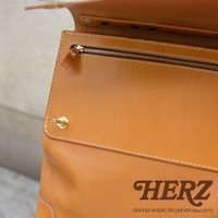 近拍職人手作皮革旅行商務兩用波士頓包，展示其光滑的淺棕色皮革材質、金色拉鍊、金屬旋轉鎖扣及縫線細節。包上印有「HERZ」品牌字樣及「HAND MADE IN JAPAN Since1973」標誌。